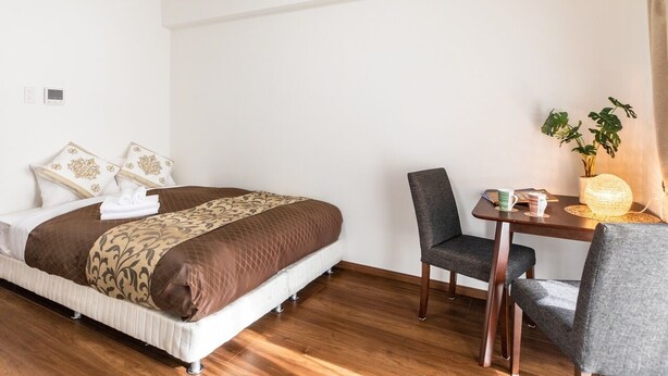A hotel room with a bed, a table with two chairs, a potted plant, and a wall-mounted thermostat. - Hotel T-style (Naha)