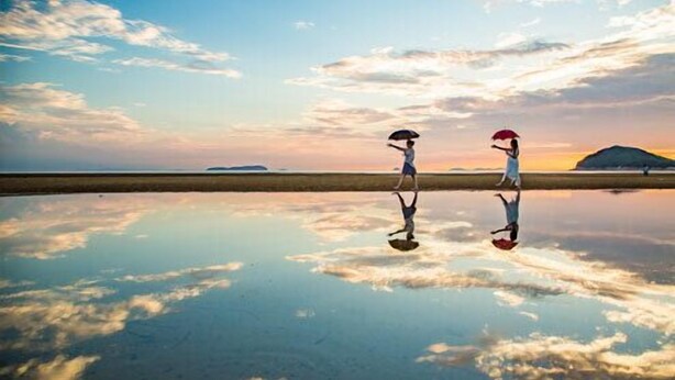 Superficie acuática reflectante con dos figuras sosteniendo paraguas, una masa de tierra distante y un cielo al atardecer.