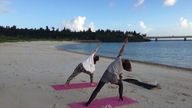 Deux personnes pratiquent le yoga sur des tapis de plage, avec une étendue d’eau et un pont en arrière-plan.