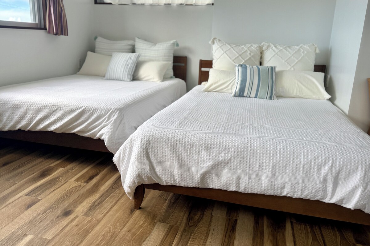 Two single beds with wooden frames and white bedding, a wooden floor, and a window with curtains.