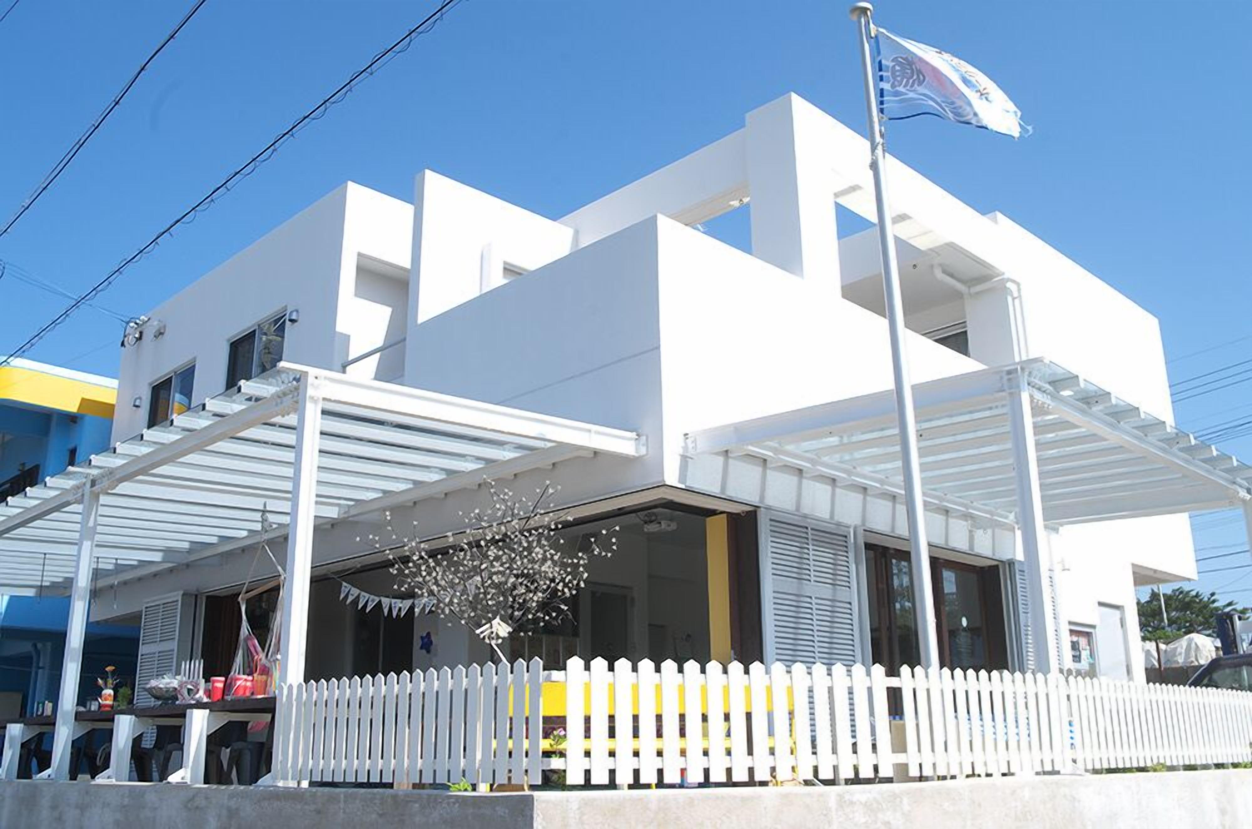 Um edifício moderno branco com alpendre coberto, mastro de bandeira e cerca de piquete branca.