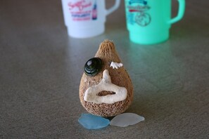 A textured object resembling a coconut with a black eye and white facial features, placed on a surface with a teal mug in the background. - Umiyado Gohan Pork Luncheon Meat (Miyakojima)