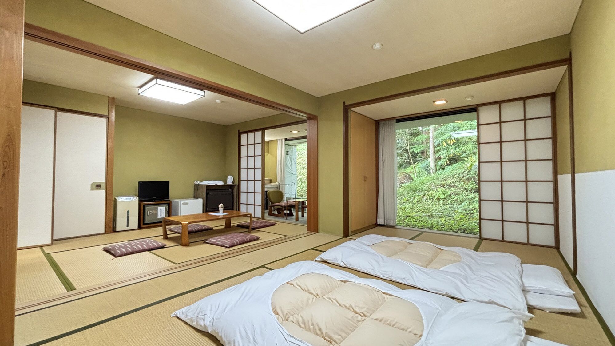 A traditional Japanese-style room with tatami flooring, a low table with cushions, a television, and a view of greenery outside.