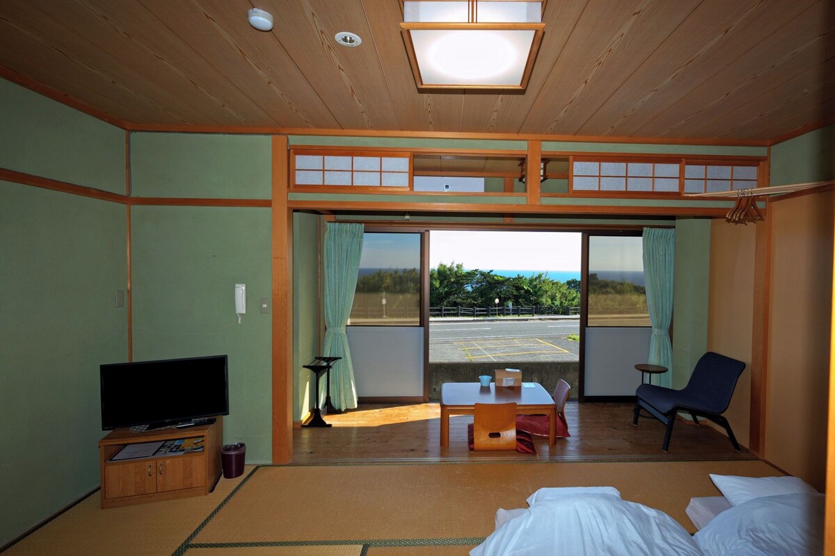 A room with a television, a desk, a chair, a bed, and a view of the outdoors.