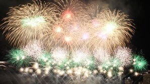 Ein Nachtbild zeigt Feuerwerk über einem Gewässer, im Hintergrund ein Steg und Gebäude.
