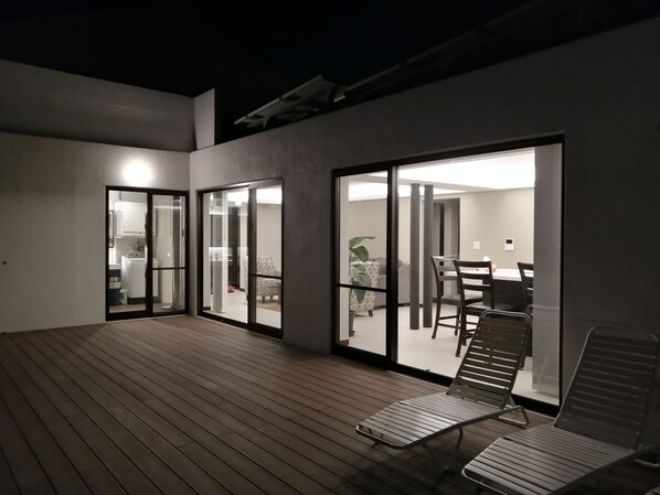A modern outdoor deck with a wooden floor, a dining area with a table and chairs, and a sliding glass door leading to a bedroom. - Salty House (Urasoe)