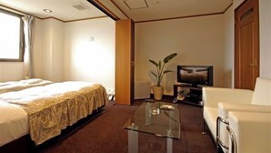Ein Hotelzimmer mit zwei Betten, einem Fernseher, einem Glastisch, einem weißen Sessel und einer Zimmerpflanze.