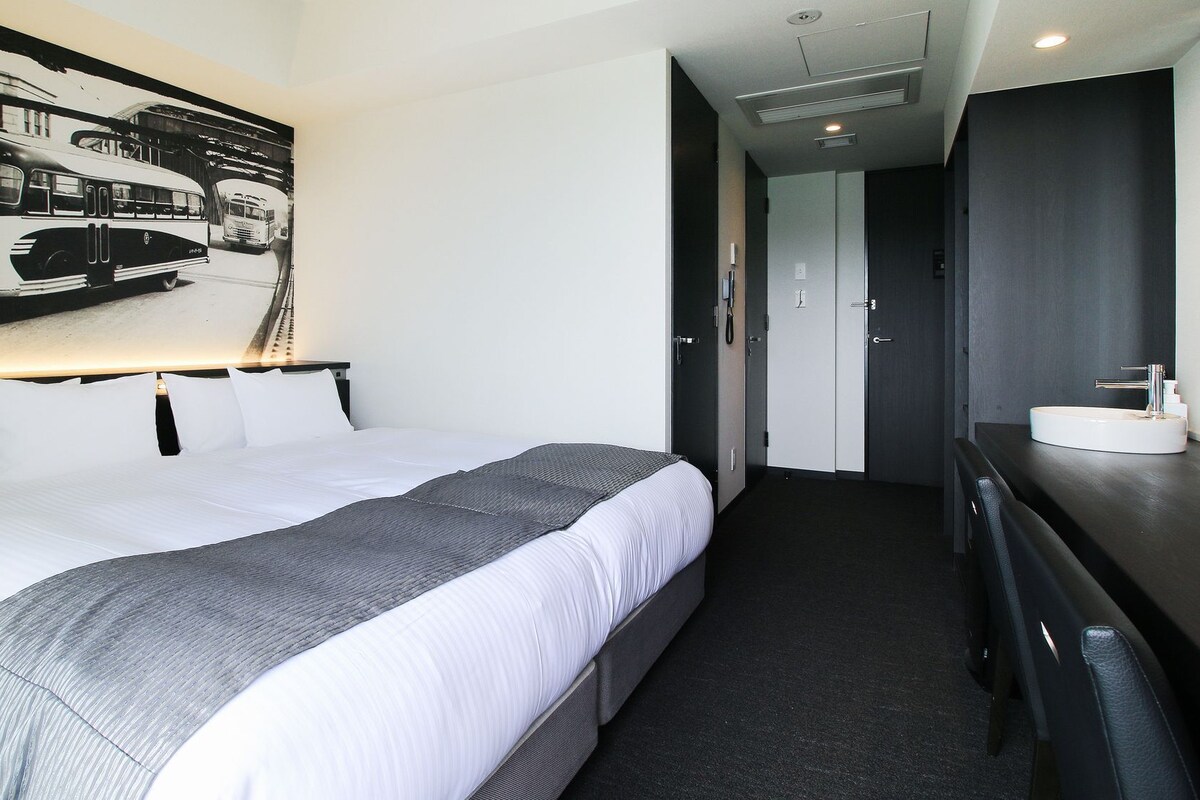 A hotel room with a large bed, a bedside table with a mirror, a wall-mounted artwork, and a separate bathroom with a sink and toilet.