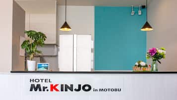A reception desk at HOTEL Mr. KINJO in MOTOBU with a sign, a potted plant, and a welcome sign with flowers.