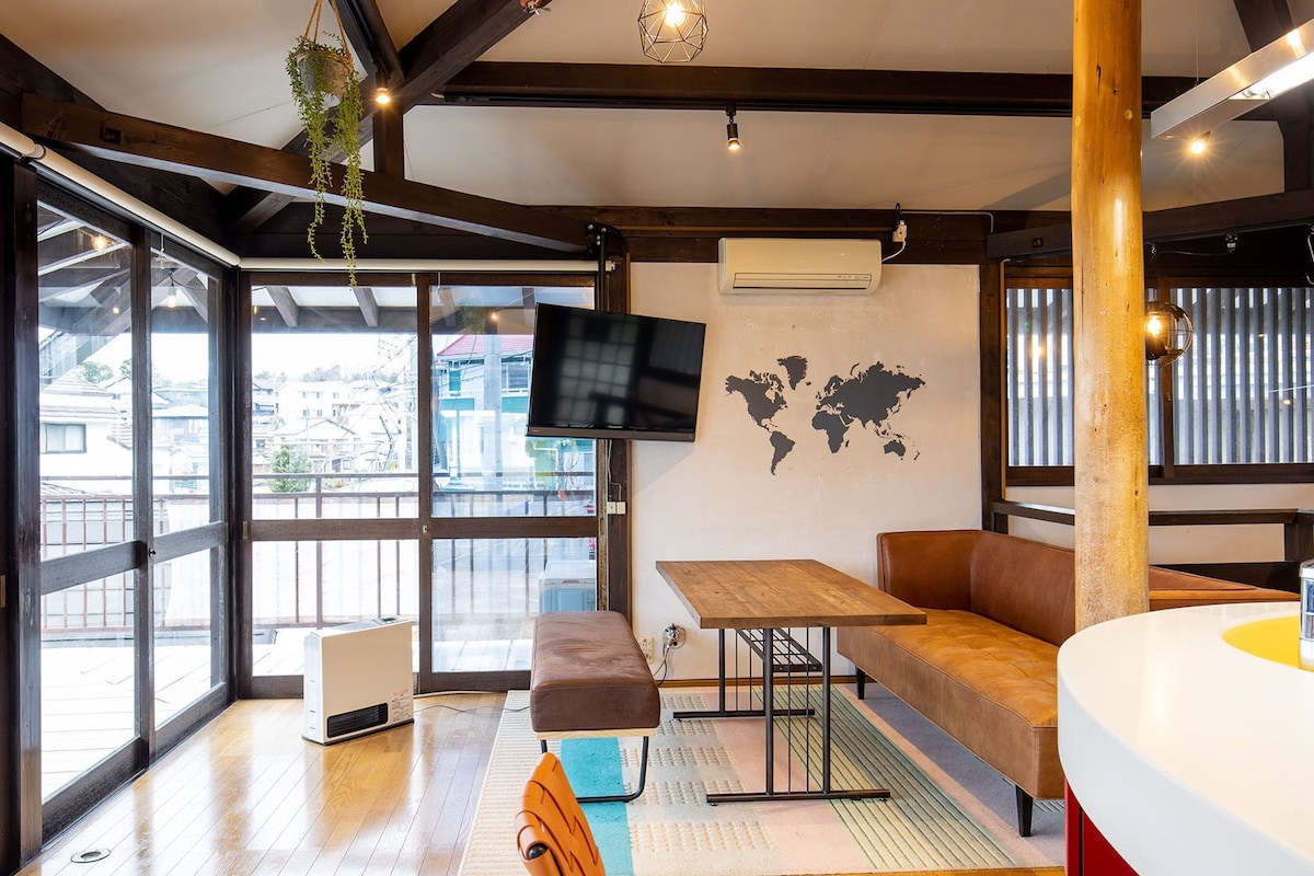 Un interior modern amb grans finestres, un televisió de pantalla plana, un mapa mundial a la paret, una taula de fusta, una banca i un bany amb rentaplatets groc.