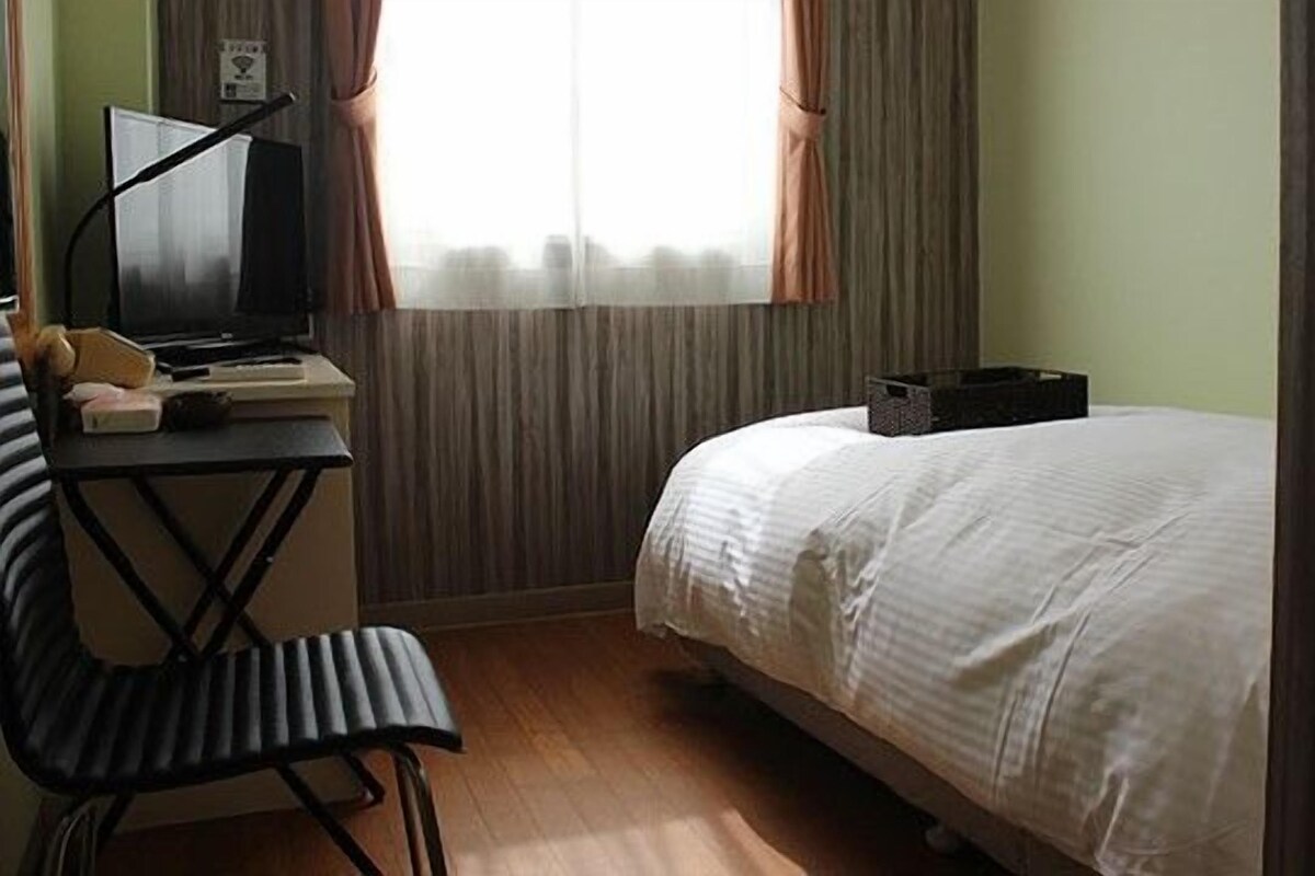 A hotel room with a bed, a desk with a chair, a television, and a window with curtains.