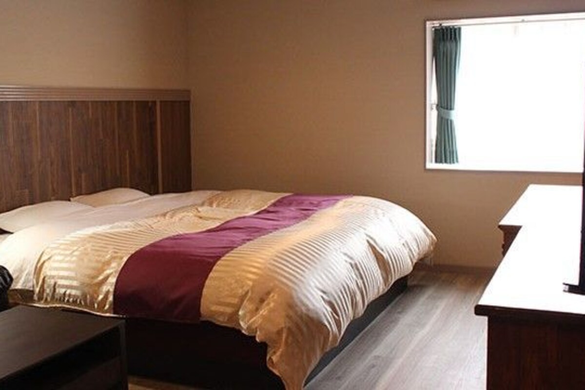 A hotel room with a bed, a wooden headboard, a window with curtains, a desk, and a television.