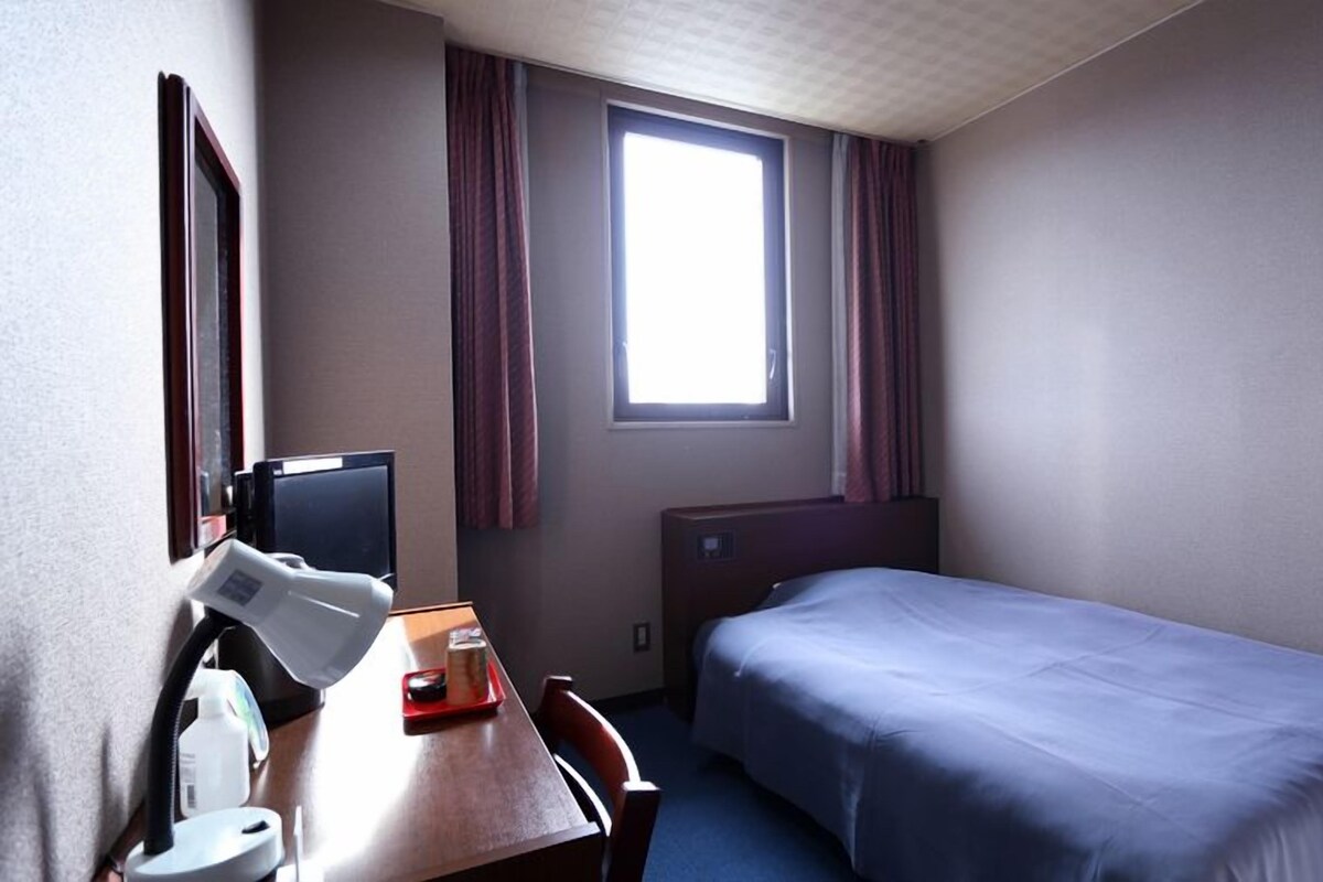 A hotel room with a single bed, a desk with a chair, a TV, a window with curtains, and a small table with a tray.
