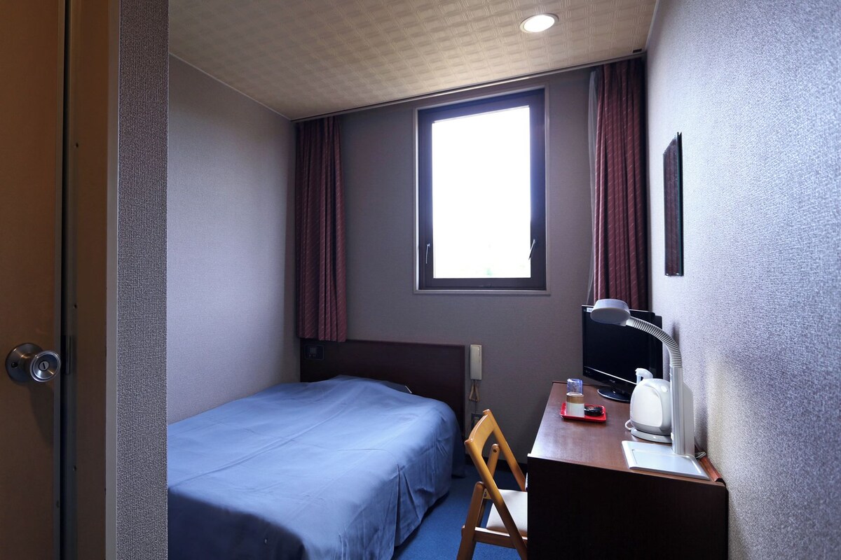 A small hotel room with a single bed, a desk with a chair, a television, and a window with curtains.