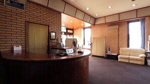 Zona de recepción con un mostrador de madera, un calendario, un cuadro enmarco y un área de descanso con un sofá y cortinas.