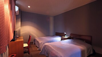 Habitación de hotel con dos camas, un escritorio con silla, una mesita con lámpara y aire acondicionado instalado en la pared.