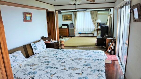 A bedroom with a large bed, wooden headboard, a bedside table, a dresser, a television, and a sliding glass door leading to a balcony.