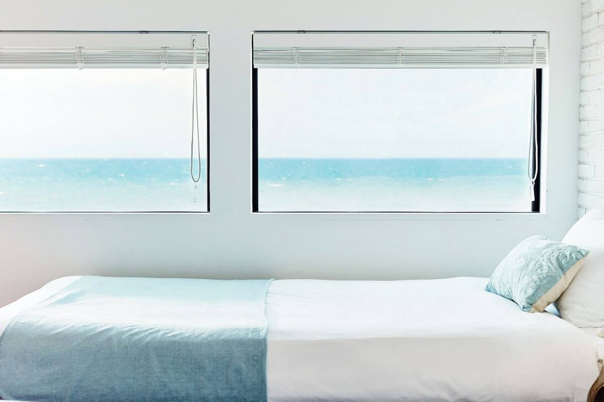 A bedroom with a bed, two windows with blinds, and a view of the sea.