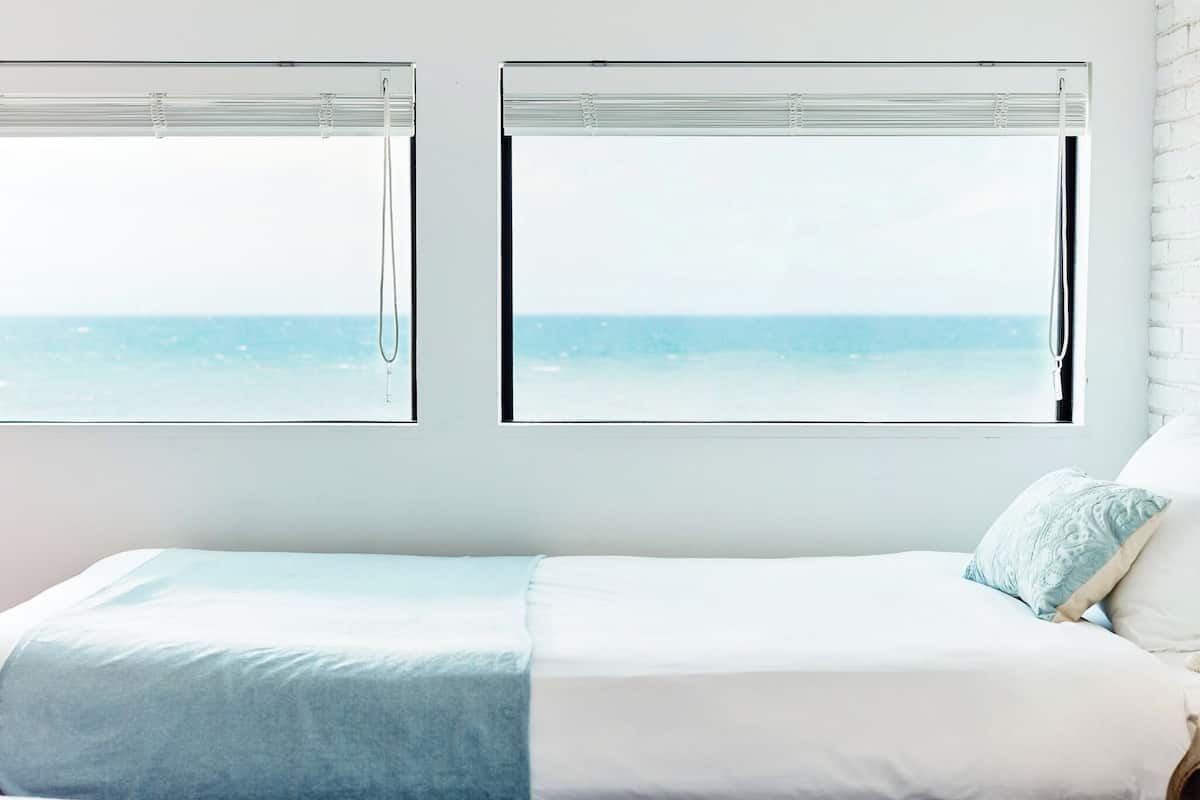 A bedroom with a bed, two windows with blinds, and a view of the sea.