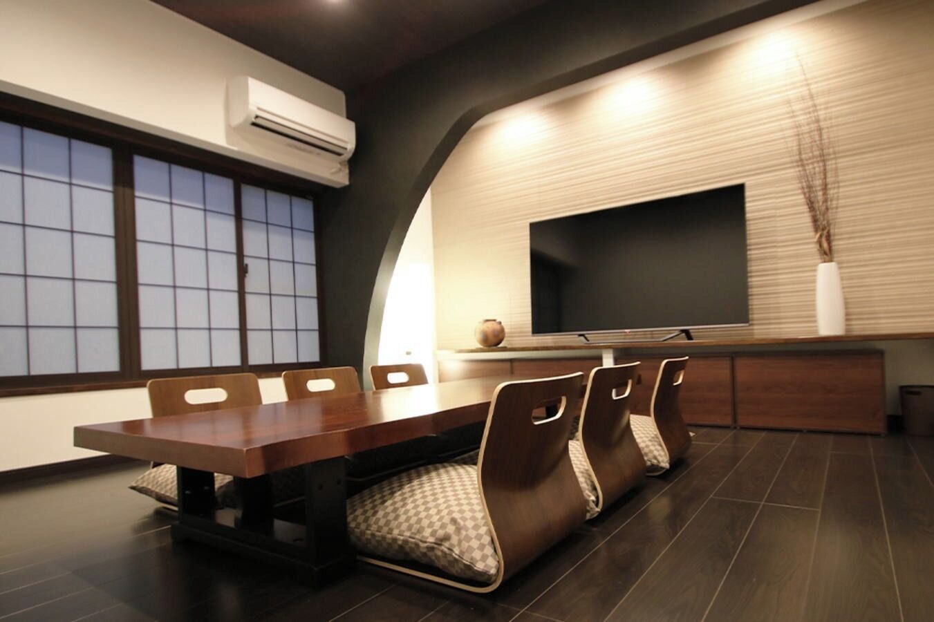 A modern dining area with a long table, cushioned chairs, a flat-screen TV, and a decorative vase.