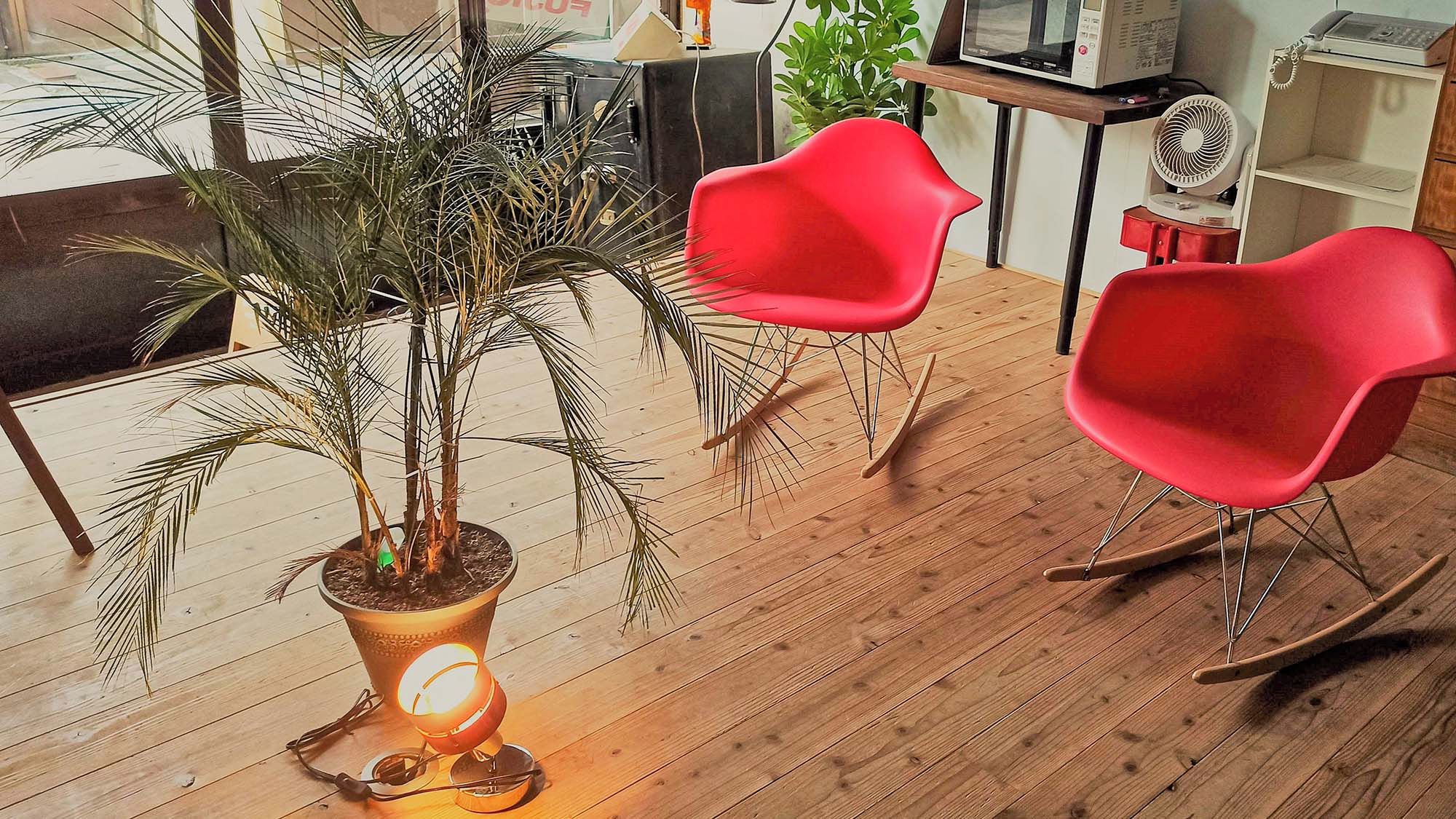 A room with a wooden floor, two red chairs, a potted plant, and a table with a lamp.