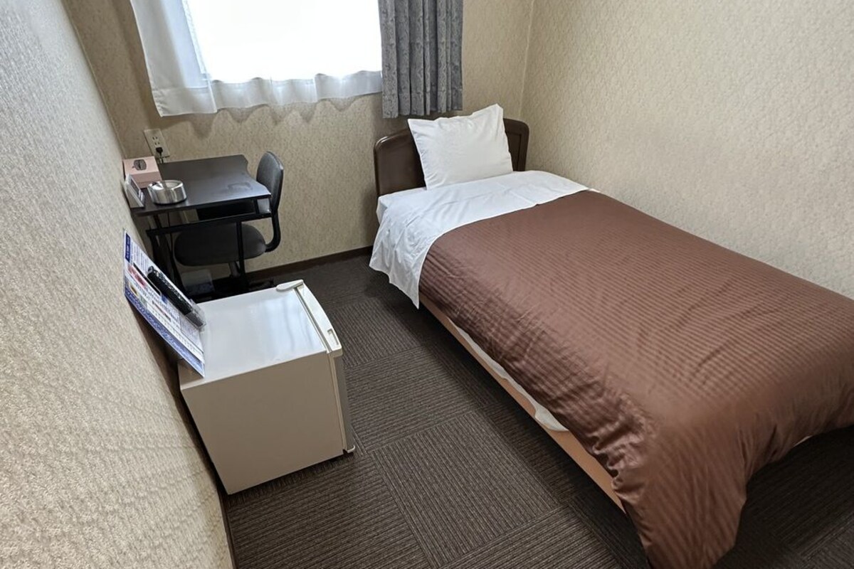A hotel room with a single bed, a desk with a chair, a small table, a drawer unit, and a pair of shoes on the floor.