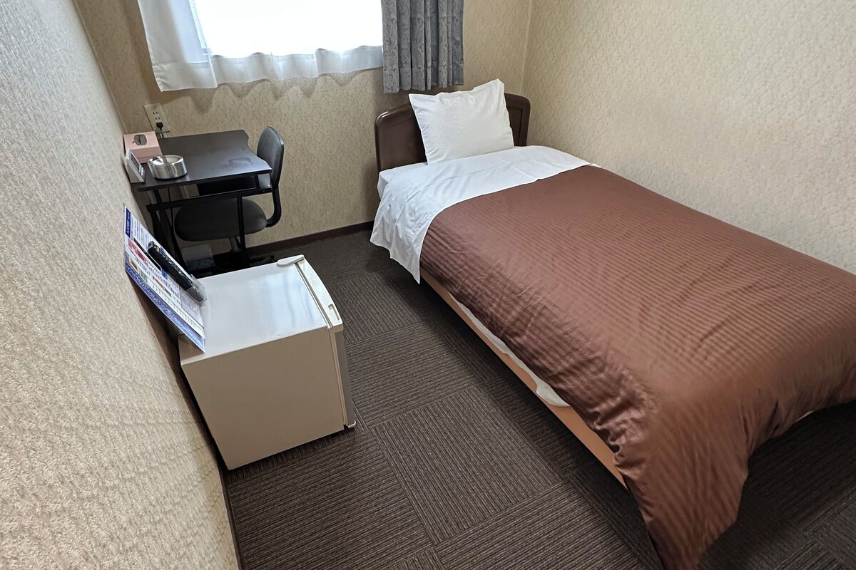 A hotel room with a bed, a desk, a chair, a shoe rack, and a window with curtains.