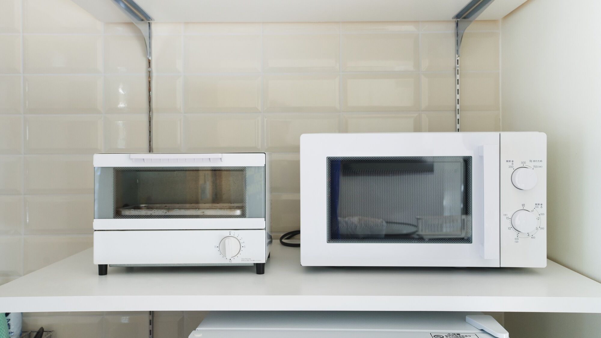 Two white microwave ovens on a white shelf against a tiled wall.