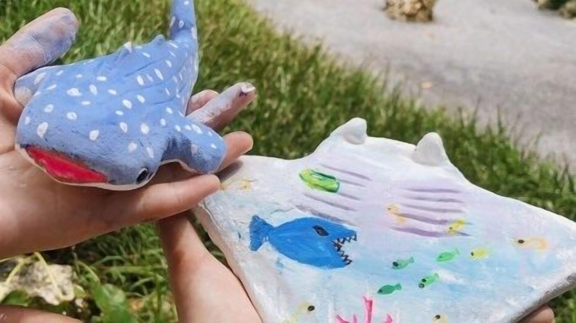 Dos esculturas de tiburón pintadas a mano, una azul y otra blanca, con un pez y coral pintados en la blanca.