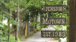 Un cartello in legno con su scritto 'PENSION MOTOWN EST. 1989', sullo sfondo di una strada alberata.