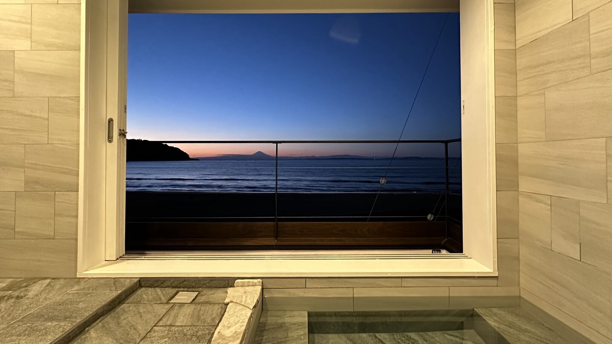 A room with a large window offering a view of the sea and a distant mountain at sunset.