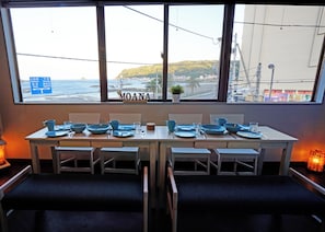Ein Esstisch gedeckt mit blauem Geschirr, Stühle und ein Fenster mit Blick auf das Meer und die Hügel.