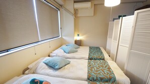 A hotel room with two beds, each with a patterned bedspread and pillows. There is a window with blinds, a wall-mounted air conditioner, and a nightstand with a lamp. - Izu no Ie Moana (Ito)
