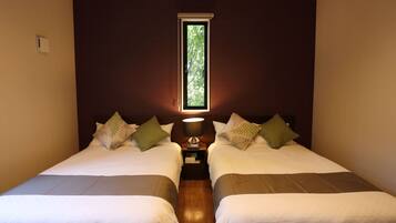 A room with two beds, a window with a view of greenery, and a bedside table with a lamp.