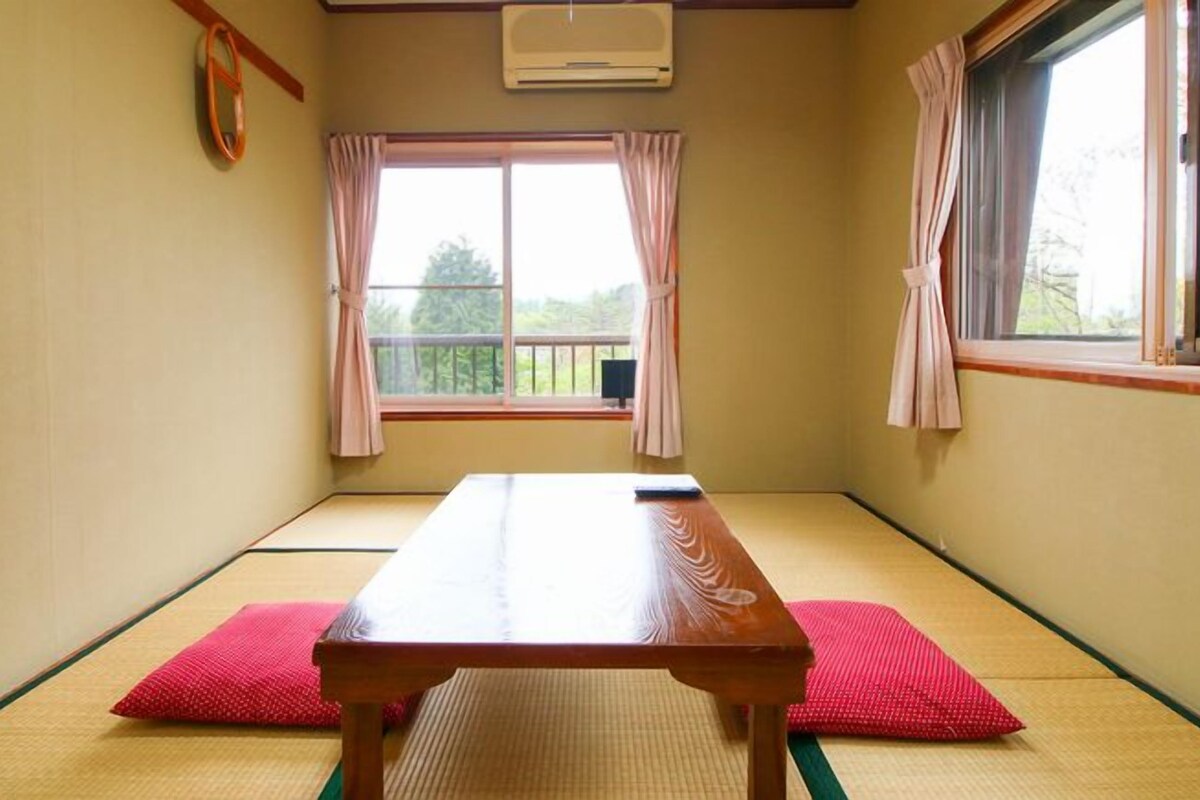 A tatami mat room with a wooden table and red cushions, large windows with curtains, and an air conditioning unit.