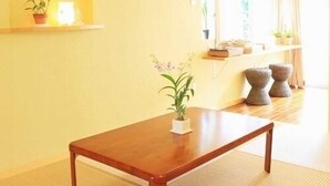 Une pièce avec une table en bois, un sol recouvert de tatamis, une étagère avec des plantes et une fenêtre avec des rideaux.