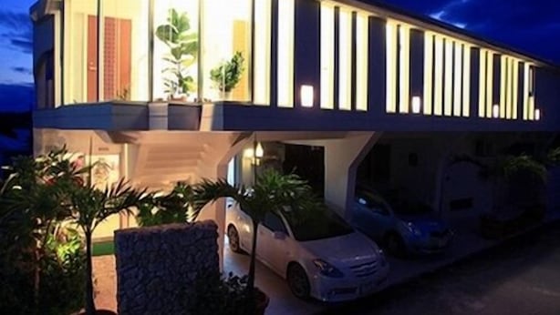 Une maison moderne à deux étages, dotée d’un toit plat, de grandes fenêtres et d’une allée où sont garées deux voitures.