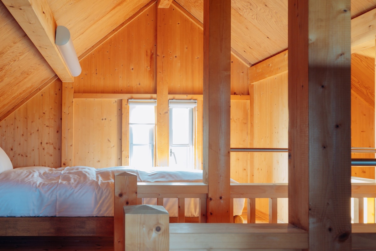 A wooden cabin with a loft-style bedroom featuring a bed, a window, and a wooden railing.