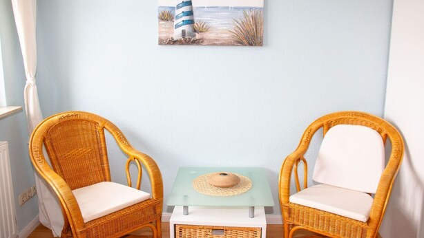 Ein Zimmer mit zwei Korbstühlen, einem kleinen Tisch mit einem Hut und einem Gemälde eines Leuchtturms an der Wand.