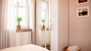 Ein Schlafzimmer mit einem Bett, einem Fenster mit weißen Vorhängen, einem Spiegel, einem Heizkörper und zwei gerahmten Bildern an der Wand.