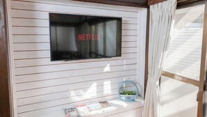 Una habitación con una televisión de pared mostrando el logo de Netflix, una mesita con revistas y una ventana con cortinas.