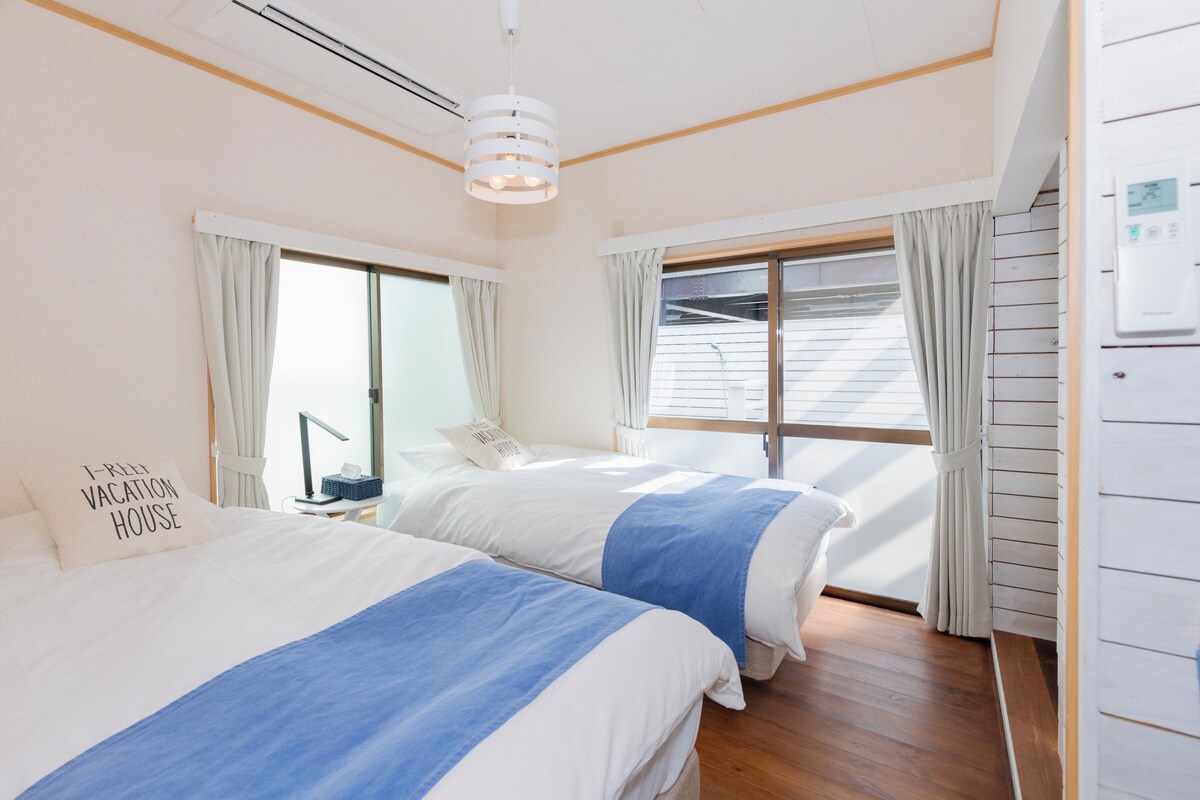 Ein Zimmer mit zwei Betten, einem Vorhangfenster, einer Deckenleuchte und einem Wandthermostat.