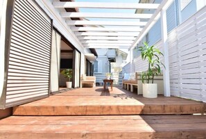 Een overdekt terras buiten met houten vloer, een zithoek met tafel en stoelen, en planten in potten.
