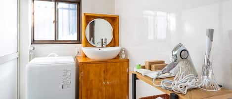 A compact bathroom with a white sink, wooden cabinet, and a mirror above it. There's a hairdryer on a wooden table, and a basket with towels.