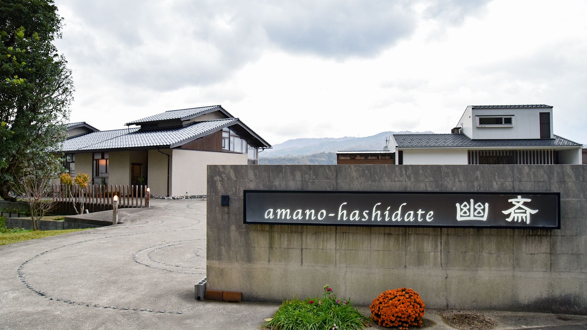 Una puerta de entrada de hormigón con un letrero que dice 'amano-hashidate' y arquitectura japonesa tradicional al fondo.