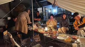Un groupe de personnes s’est réuni autour d’une table où étaient disposés de la nourriture et des boissons, à l’intérieur d’une structure ressemblant à une tente, d’où l’on pouvait apercevoir des bâtiments en arrière-plan.