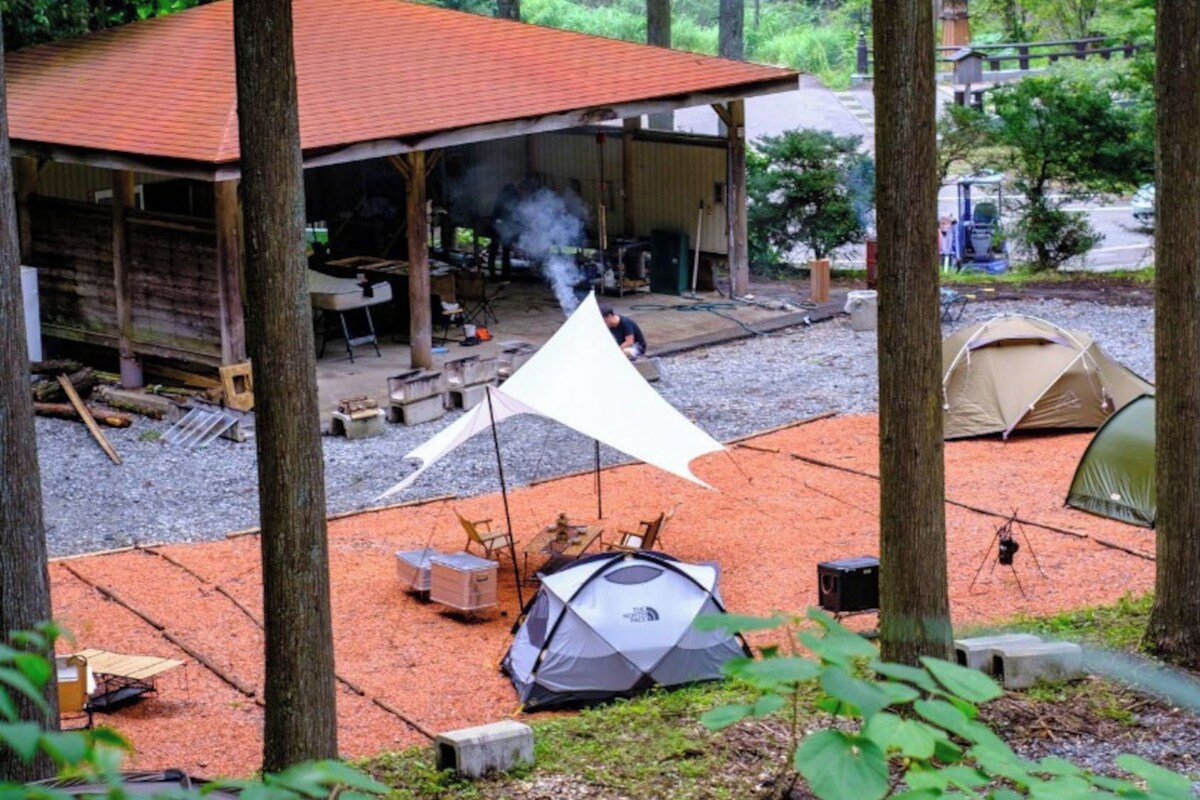 A rustic cabin with a red roof, a covered outdoor area with a table and chairs, and several tents set up on a gravel area surrounded by trees.