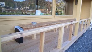 Una terraza de madera con un banco y un contenedor de basura, con vistas a una zona panorámica con árboles y un edificio al fondo.