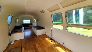 Un interior compacto y bien iluminado con piso de madera, un pequeño rincón de descanso y amplias ventanas que ofrecen luz natural y vistas al follaje.