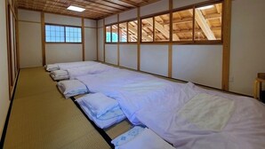 Una habitación japonesa tradicional con suelo de tatami, una cama baja con ropa de cama blanca y vigas de madera en el techo.